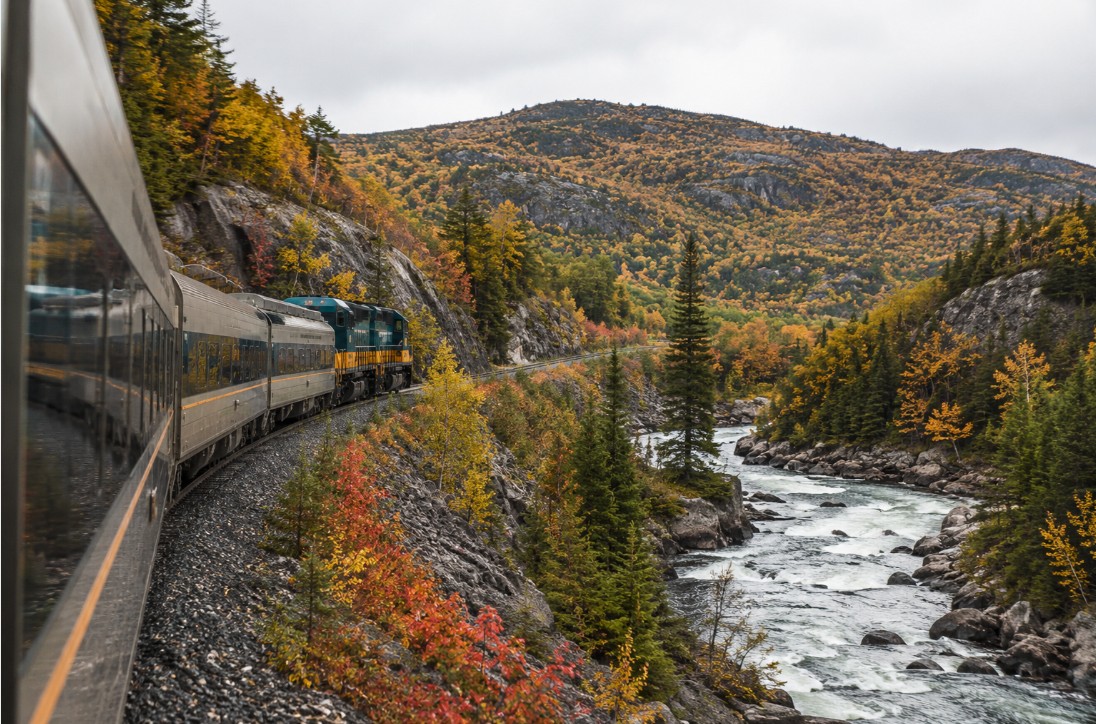 Passenger train winding through rugged Algoma forest and granite hills beside a river
